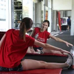 A woman in a red Crossjoin t-shirt sits on a gym mat stretching her leg while looking at her smiling reflection in a large wall mirror.