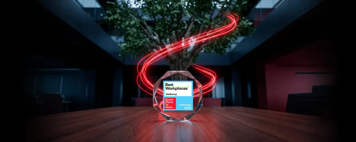 A glass octagonal award for "Best Workplaces Wellbeing, Great Place To Work, Portugal 2025" sits on a wooden table. In the background, glowing red light trails swirl around a large indoor tree within a dark, modern office space.