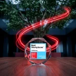A glass octagonal award for "Best Workplaces Wellbeing, Great Place To Work, Portugal 2025" sits on a wooden table. In the background, glowing red light trails swirl around a large indoor tree within a dark, modern office space.