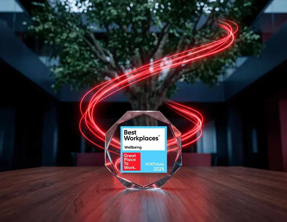 A glass octagonal award for "Best Workplaces Wellbeing, Great Place To Work, Portugal 2025" sits on a wooden table. In the background, glowing red light trails swirl around a large indoor tree within a dark, modern office space.