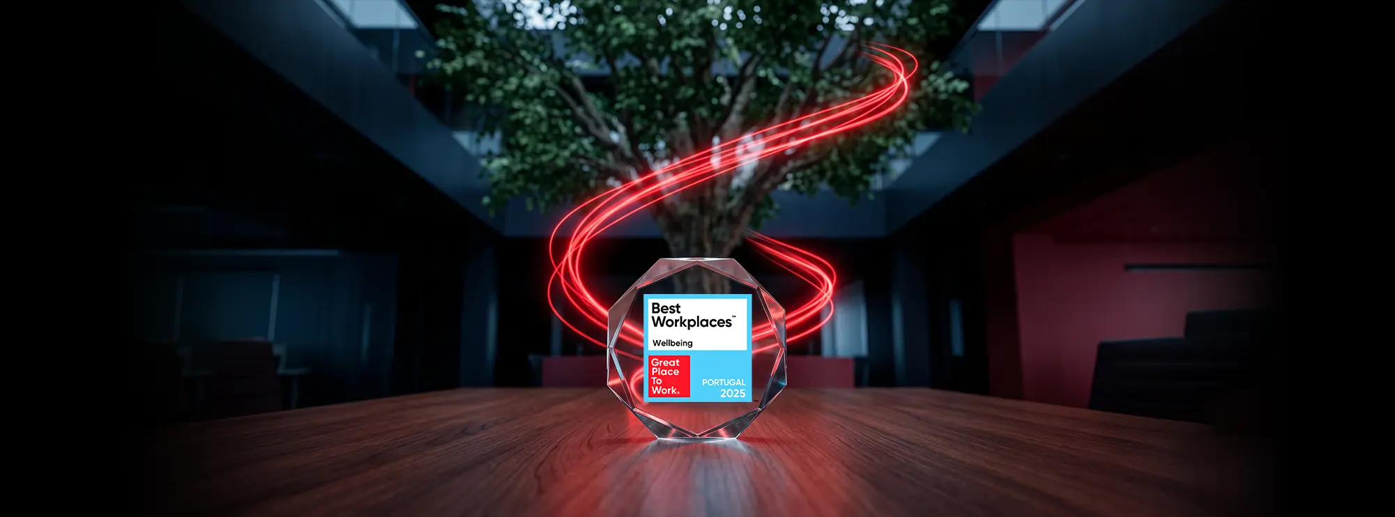 A glass octagonal award for "Best Workplaces Wellbeing, Great Place To Work, Portugal 2025" sits on a wooden table. In the background, glowing red light trails swirl around a large indoor tree within a dark, modern office space.