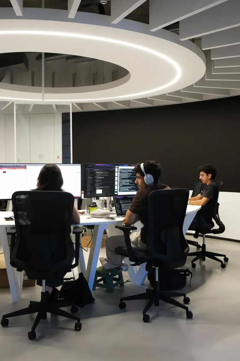 Team of young tech professionals working together at a curved desk with multiple monitors inside a modern, open-plan Crossjoin Solutions office.
