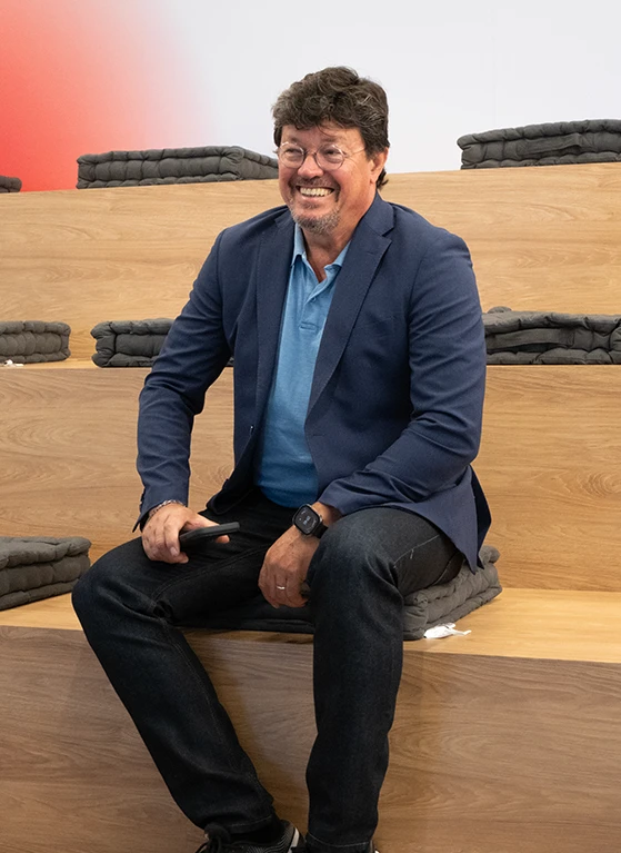 João Modesto, Co-founder and CEO of Crossjoin Solutions, smiling during an interview on leadership transition and corporate legacy.Natural and candid portrait of João Modesto, Co-founder and CEO of Crossjoin Solutions, wearing glasses, a navy blue blazer, and a blue polo shirt, visibly relaxed and smiling. He is seated on tiered wooden benches with dark cushions in a modern auditorium setting, with a softly blurred background featuring a subtle red accent. Ideal for articles, interviews, case studies, and corporate web pages focused on leadership, organizational culture, and the "Perform to Perfection" vision.