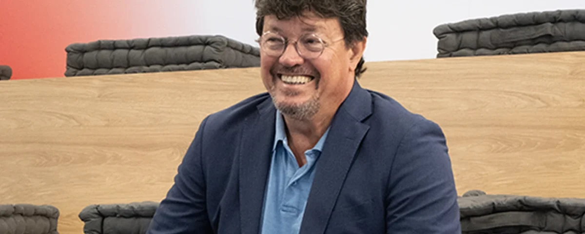 João Modesto, Co-founder and CEO of Crossjoin Solutions, smiling during an interview on leadership transition and corporate legacy.Natural and candid portrait of João Modesto, Co-founder and CEO of Crossjoin Solutions, wearing glasses, a navy blue blazer, and a blue polo shirt, visibly relaxed and smiling. He is seated on tiered wooden benches with dark cushions in a modern auditorium setting, with a softly blurred background featuring a subtle red accent. Ideal for articles, interviews, case studies, and corporate web pages focused on leadership, organizational culture, and the "Perform to Perfection" vision.