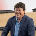 João Modesto, Co-founder and CEO of Crossjoin Solutions, smiling during an interview on leadership transition and corporate legacy.Natural and candid portrait of João Modesto, Co-founder and CEO of Crossjoin Solutions, wearing glasses, a navy blue blazer, and a blue polo shirt, visibly relaxed and smiling. He is seated on tiered wooden benches with dark cushions in a modern auditorium setting, with a softly blurred background featuring a subtle red accent. Ideal for articles, interviews, case studies, and corporate web pages focused on leadership, organizational culture, and the "Perform to Perfection" vision.