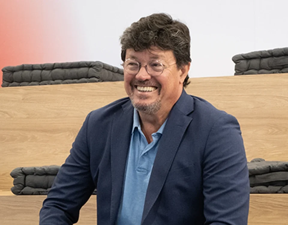 João Modesto, Co-founder and CEO of Crossjoin Solutions, smiling during an interview on leadership transition and corporate legacy.Natural and candid portrait of João Modesto, Co-founder and CEO of Crossjoin Solutions, wearing glasses, a navy blue blazer, and a blue polo shirt, visibly relaxed and smiling. He is seated on tiered wooden benches with dark cushions in a modern auditorium setting, with a softly blurred background featuring a subtle red accent. Ideal for articles, interviews, case studies, and corporate web pages focused on leadership, organizational culture, and the "Perform to Perfection" vision.