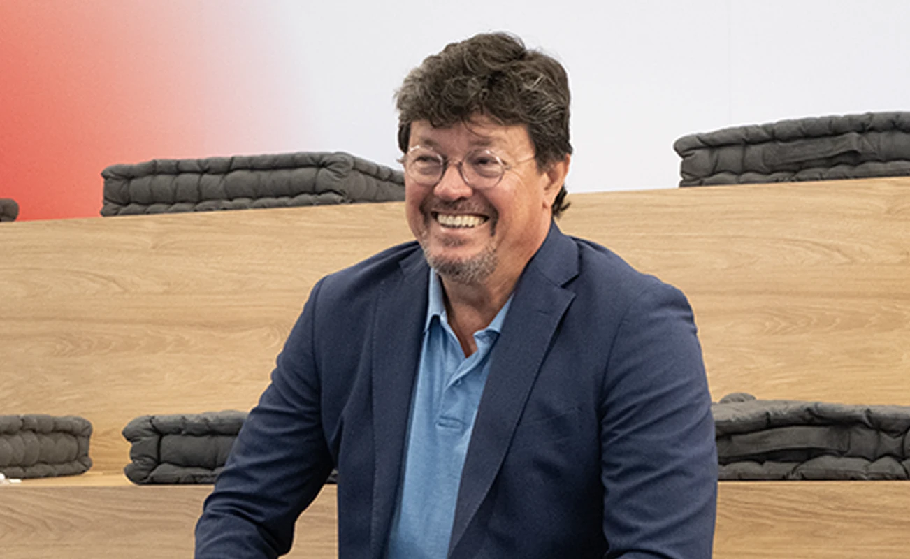 João Modesto, Co-founder and CEO of Crossjoin Solutions, smiling during an interview on leadership transition and corporate legacy.Natural and candid portrait of João Modesto, Co-founder and CEO of Crossjoin Solutions, wearing glasses, a navy blue blazer, and a blue polo shirt, visibly relaxed and smiling. He is seated on tiered wooden benches with dark cushions in a modern auditorium setting, with a softly blurred background featuring a subtle red accent. Ideal for articles, interviews, case studies, and corporate web pages focused on leadership, organizational culture, and the "Perform to Perfection" vision.